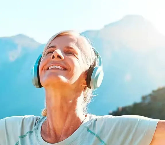 Woman with headphones smiling with her face turned toward the sun, mountains behind her